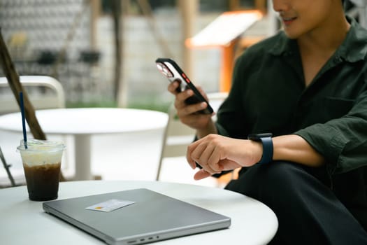 Smiling man relaxes at an outdoor cafe and using smartphone beside a closed laptop.