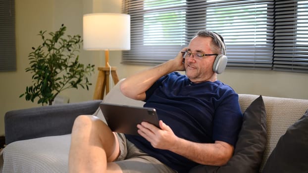 Senior man wearing headphones relaxing on the couch and using a digital tablet.