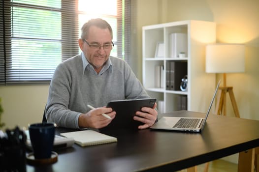 Senior man in sweater and glasses working from home using digital tablet.