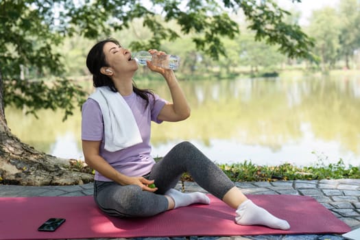 A woman takes a refreshing sip of water while sitting on a yoga mat by the serene lake, promoting wellness and hydration.