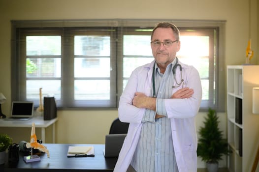 Portrait of experienced male physician with arms crossed in a bright workspace.