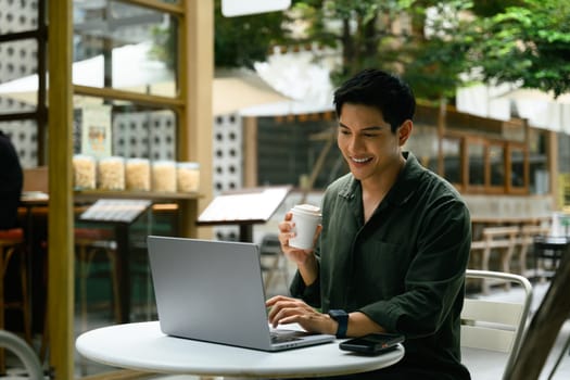 Handsome male freelancer using laptop and sipping coffee in a relaxed outdoor workspace.
