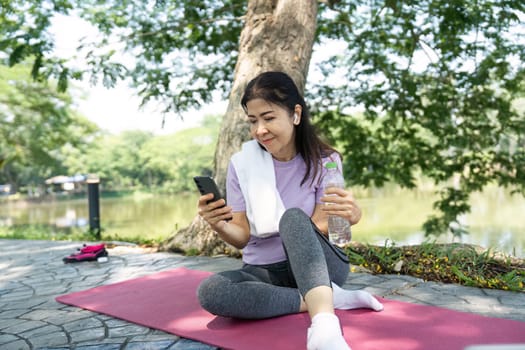 A relaxed woman sits on a yoga mat by the lake, checking her phone after exercising, highlighting wellness and connectivity.