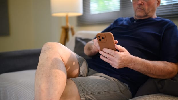 Mature male browsing or texting on mobile phone, relaxing on couch at home.