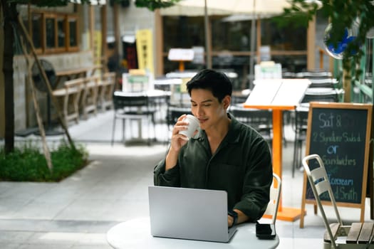 Handsome male freelancer using laptop and sipping coffee in a relaxed outdoor workspace.