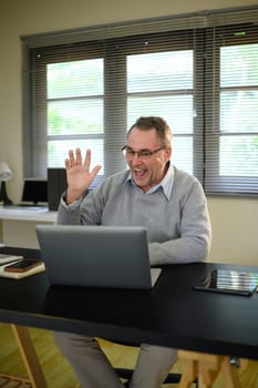 Confident older man enjoys an online conversation from his tidy home office.