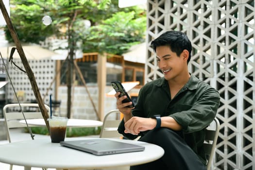 Smiling man relaxes at an outdoor cafe and using smartphone beside a closed laptop.