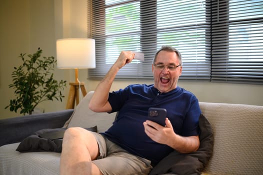 Excited senior man holding credit card and phone, joyful expression after successful transaction.