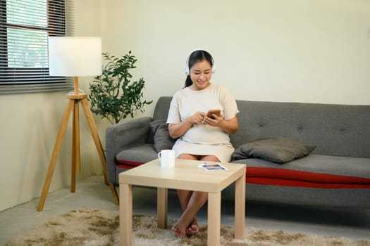 Relaxed pregnant woman sitting on sofa with headphones and using smartphone.