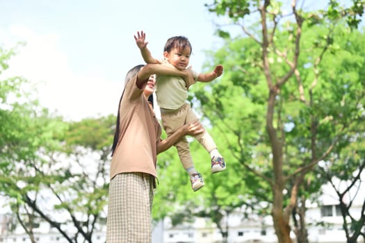 Mother smiles as she lifts her young child into the air during a sunny day at the park.