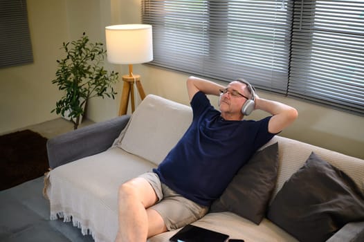 Relaxed older adult laying back on a sofa, listening to music with headphones.