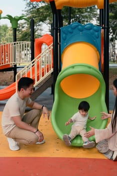 Happy parents watch and cheer as their toddler slides down a colorful slide.