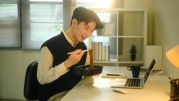 Smiling man eating instant noodles while working at his desk in a cozy, sunlit home office.