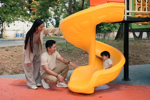 Happy parents watch and cheer as their toddler slides down a colorful slide.