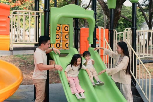 Parents cheer joyfully as two young children enjoy sliding on a playground slide in a sunny park.