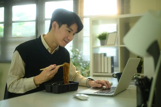 Smiling man eating instant noodles while working at his desk in a cozy, sunlit home office.