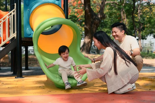 Happy parents watch and cheer as their toddler slides down a colorful slide.