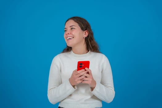 Happy young woman holding a red smartphone while looking to the side with a surprised expression. She stands in front of a bright blue background, expressing excitement and curiosity.
