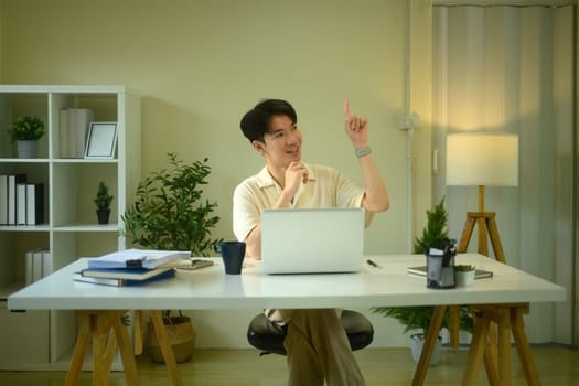 Smiling man entrepreneur sitting at working desk and pointing finger up.