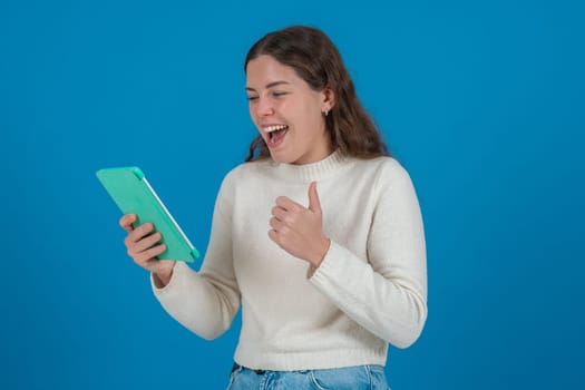 Happy young Caucasian woman holding tablet and showing thumbs up with a cheerful expression. Studio shot on blue background with copy space, ideal for communication or tech concepts.