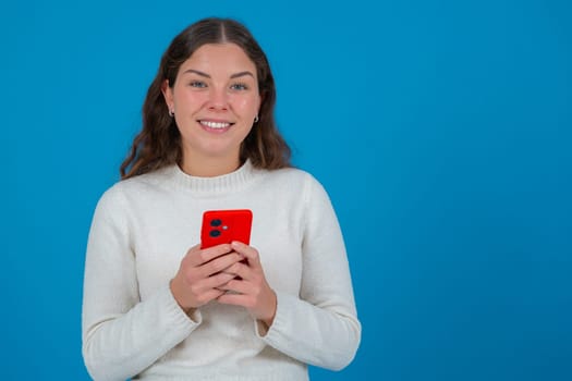Cheerful young woman in white sweater looking at camera while holding a red smartphone in her hands, standing against a vivid blue background, representing connection, joy.copy space