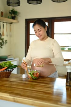 Smiling expectant woman adds dressing to a glass bowl of vegetable salad.