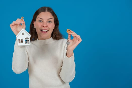 Happy young woman holding a house model and keys, expressing excitement. She wears a white sweater and stands against a blue background. Concept of home ownership, real estate, or investment. Copy space