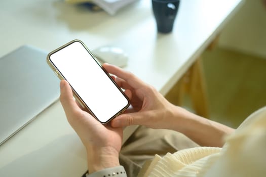 Hands holding smartphone with empty screen sitting in front of laptop at desk.