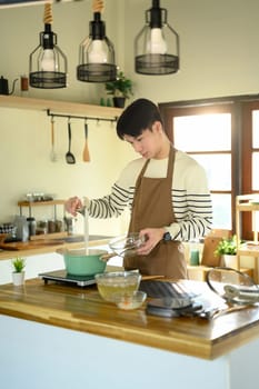 Man in apron pouring ingredients into a pot while preparing a homemade meal.