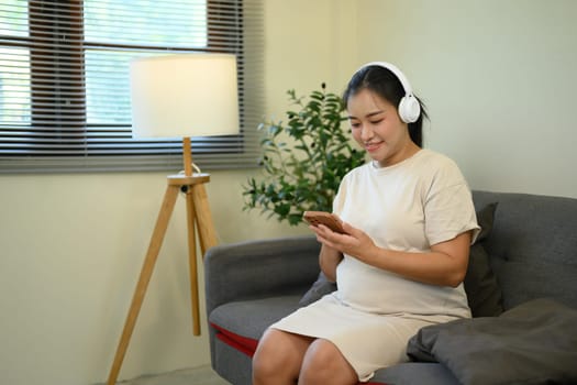 Relaxed pregnant woman sitting on sofa with headphones and using smartphone.