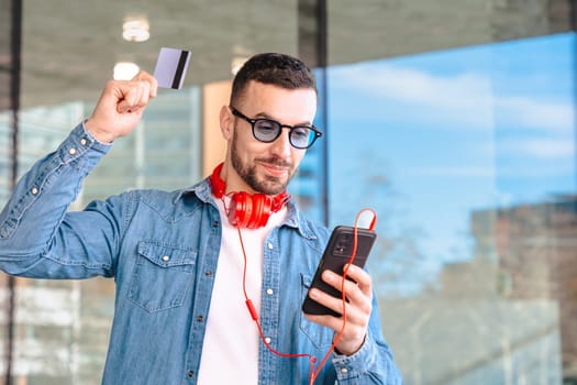 Happy man celebrating a successful online purchase while holding a credit card and smartphone. He smiles with excitement, wearing headphones and sunglasses in a modern urban environment.