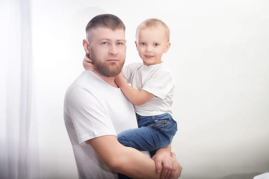A father holds his young son in his arms, both are dressed in casual outfits and smiling joyfully in a well-lit indoor setting.