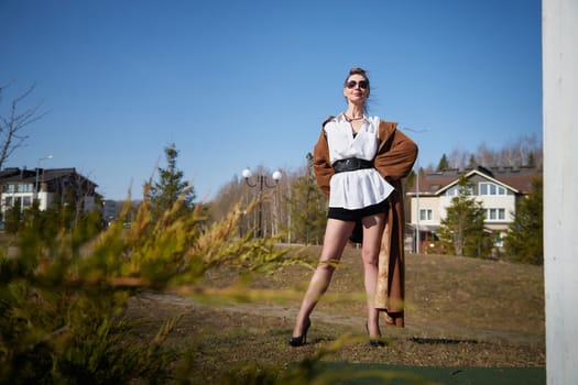 A model showcases a fashionable look with a flowing coat and high heels, standing confidently in a sunny outdoor setting.