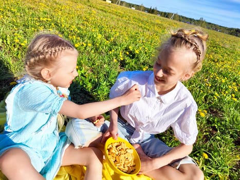 Two girls sit together in a dandelion-covered field, sharing snacks and enjoying each other's company under a clear blue sky.