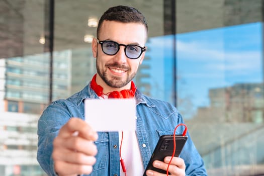 Young caucasian man smiling confidently at the camera while holding a credit card and smartphone. He stands in an urban setting with headphones around his neck, ready for a digital transaction.