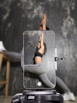 Young fitness influencer recording workout video at home using smartphone. Sporty woman in activewear doing stretching exercises for online audience during live fitness session