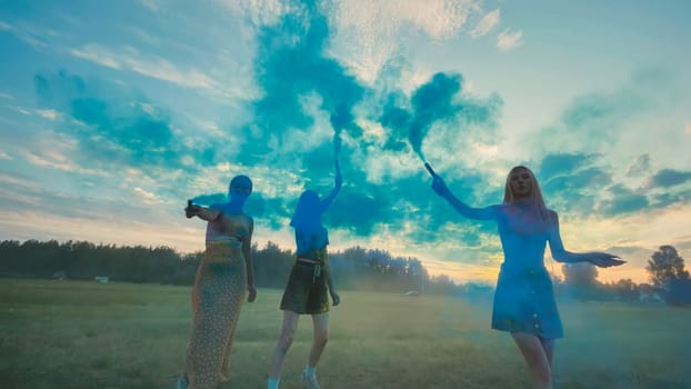 Three friends from school delight in a stunning sunset as they play with blue smoke bombs in a field, crafting a vivid and unforgettable moment