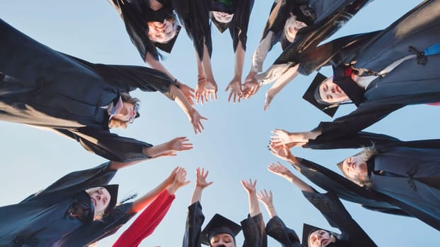 Group of students wearing graduation gowns and caps raising hands together forming circle in the sky, celebrating graduation day
