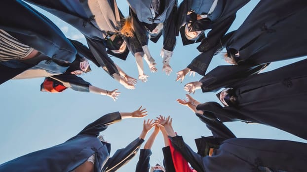 Group of students wearing graduation gowns joining hands forming a circle against blue sky, celebrating their graduation day
