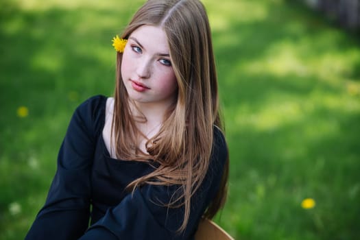 Portrait of a teenage student girl sitting outdoors with a dandelion tucked behind her ear, enjoying her school break on a grassy field