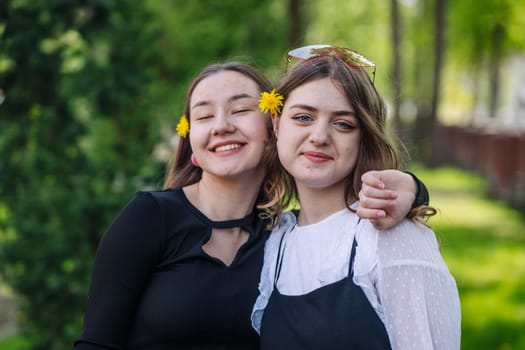 Two cheerful female students with dandelions in their hair are embracing, enjoying a pleasant moment during their school day