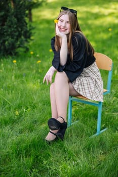 Cheerful young woman sitting on a chair in a grassy outdoor setting, holding a yellow flower, enjoying a sunny school day in nature