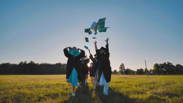 Group of cheerful graduates wearing gowns and caps, celebrating their graduation by joyfully throwing books in the air in a field at sunset