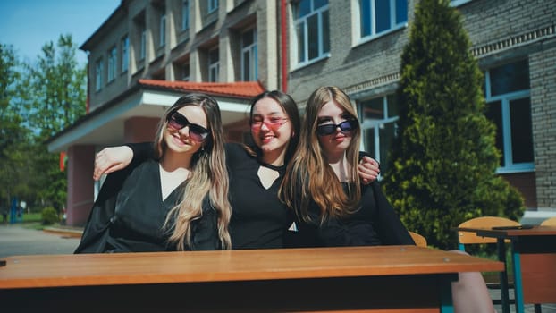Three cheerful female students wearing sunglasses are posing, embracing each other, sitting at a desk outside their school on a sunny day