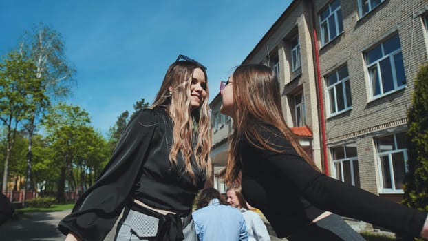 Two girls kissing each other on the cheek in front of their high school, celebrating friendship and affection