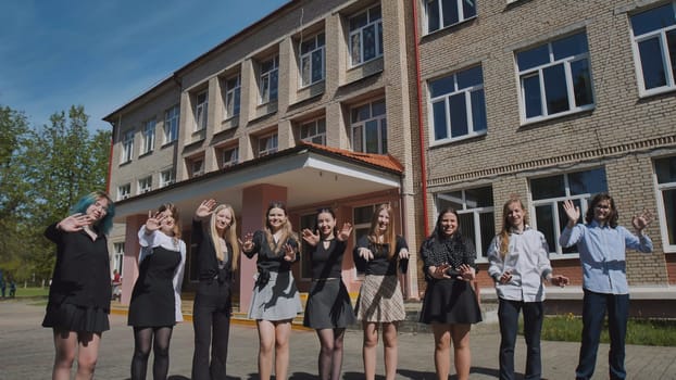 Group of cheerful students waving hands while standing together in front of school building on sunny day, celebrating end of school year
