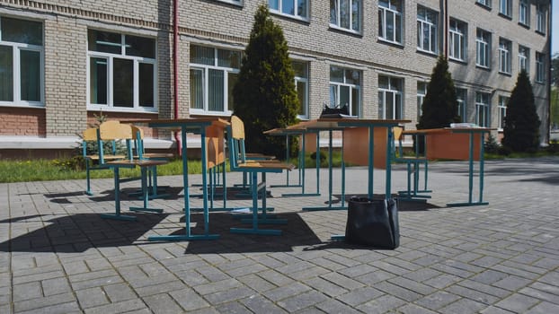 Empty school desks and chairs standing outdoors next to school building during school day with books and bag on the ground