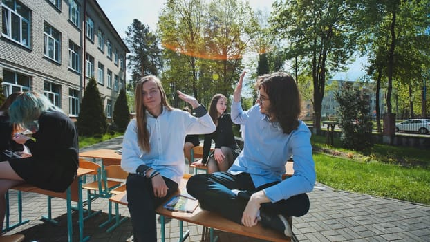 Group of happy students giving high five while sitting on school desks outdoors during break time