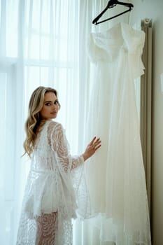 Bride's morning. gentle and charming blonde in lace robe stands near elegant wedding dress. she has long wavy hair, looks at camera. young woman in silk robe and lingerie.