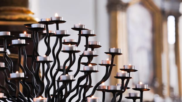Many small burning candles on an elegant wrought iron candelabrum illuminate a church interior